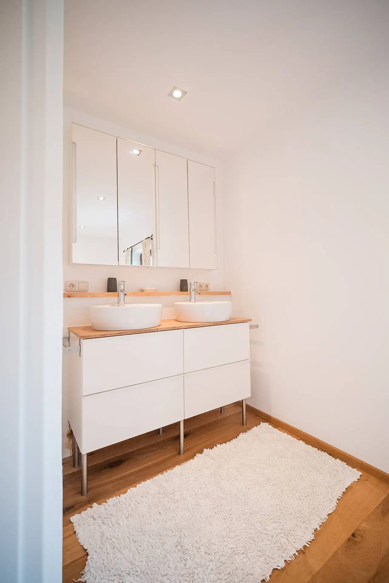 Modernes Badezimmerinterieur mit Doppelwaschbecken, großem Spiegel und einem weißen Zottelteppich auf Holzboden.