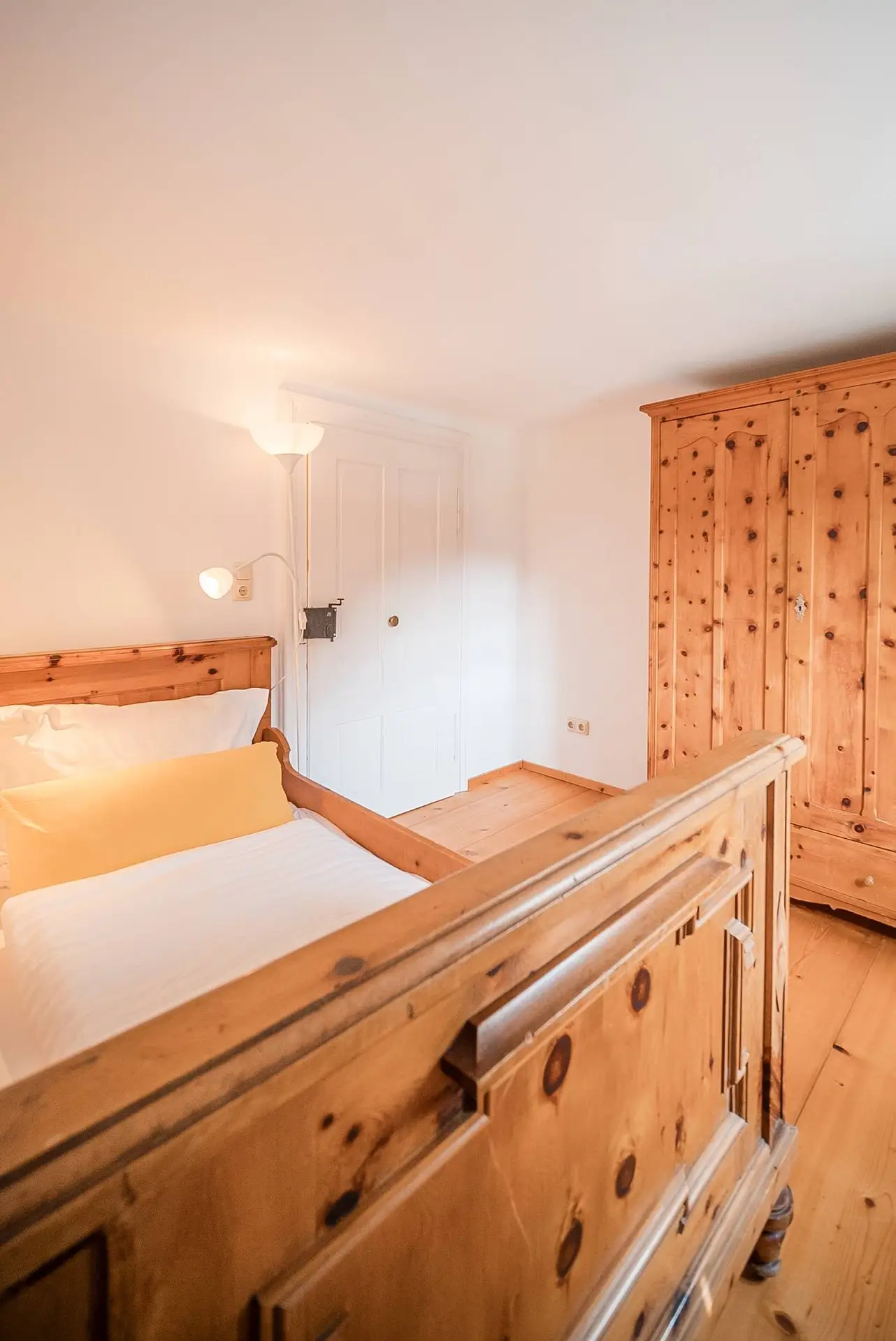 Gemütliches Schlafzimmer mit Bettgestell und Kommode aus Holz, weißer Bettwäsche und einer Wandlampe, mit weißer Tür und warmer Beleuchtung.