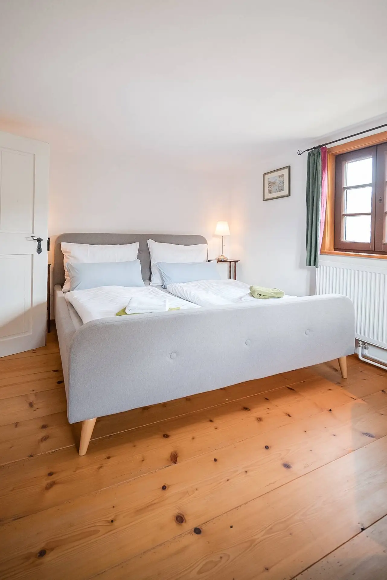 Gemütliches Schlafzimmer mit einem Doppelbett mit weißer Bettwäsche, Holzboden, einem Fenster mit Vorhängen und einer Nachttischlampe.