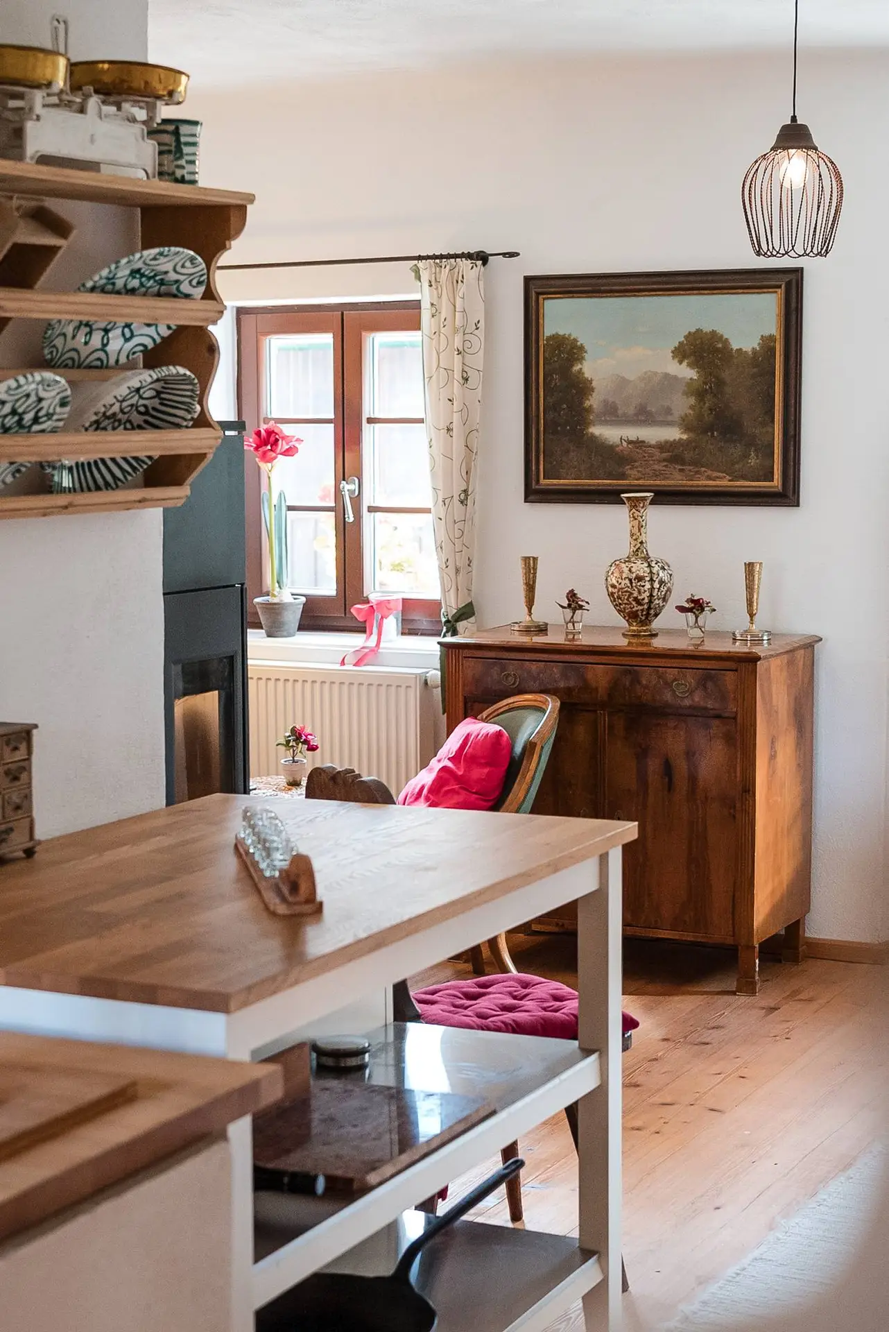 Ein gemütliches, sonnendurchflutetes Esszimmer mit einem Holztisch, Regalen mit Büchern und Dekorationsgegenständen und einer Hängelampe darüber.