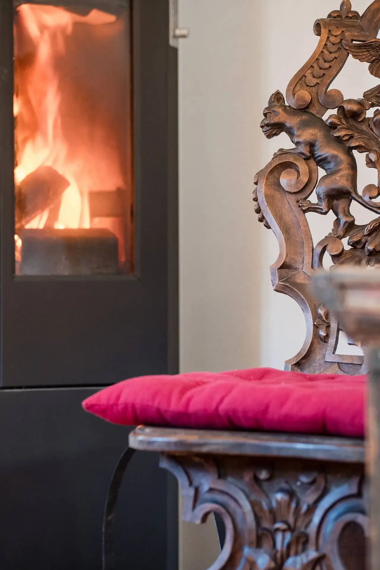 Alter, verzierter Holzstuhl mit einem rosa Kissen im Fokus, im Hintergrund ist ein brennender Kamin zu sehen.