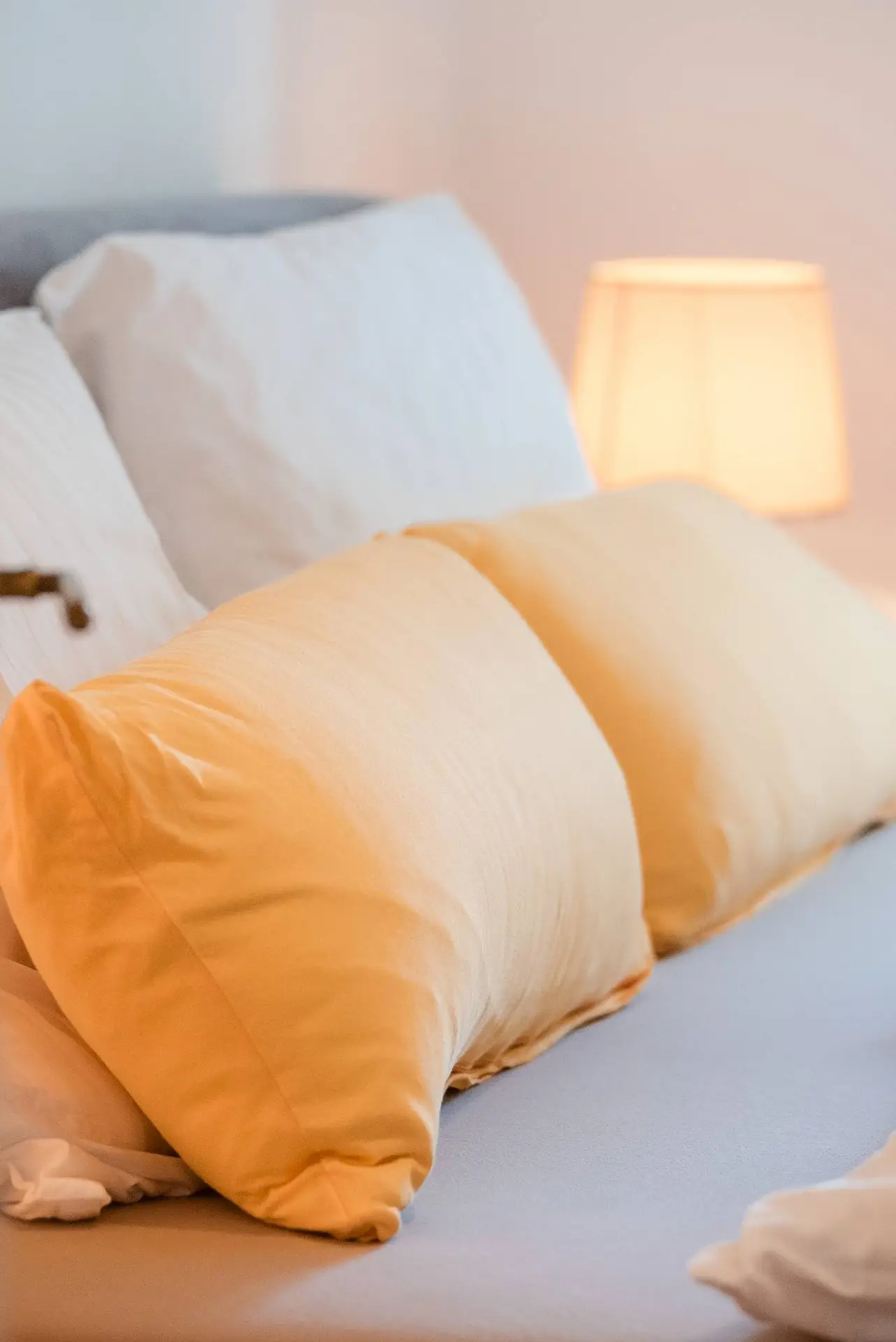 Eine gemütliche Schlafzimmerszene mit einem ordentlich gemachten Bett mit flauschigen weißen und gelben Kissen und einer sanft leuchtenden Nachttischlampe.