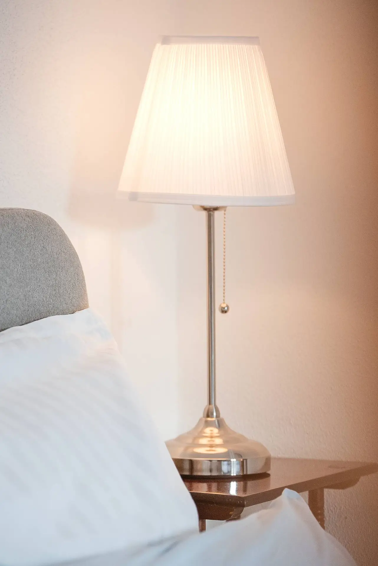 Ein Nachttisch mit einer Lampe mit weißem Schirm und Metallsockel, neben einem grau gepolsterten Kopfteil und weißen Kissen.