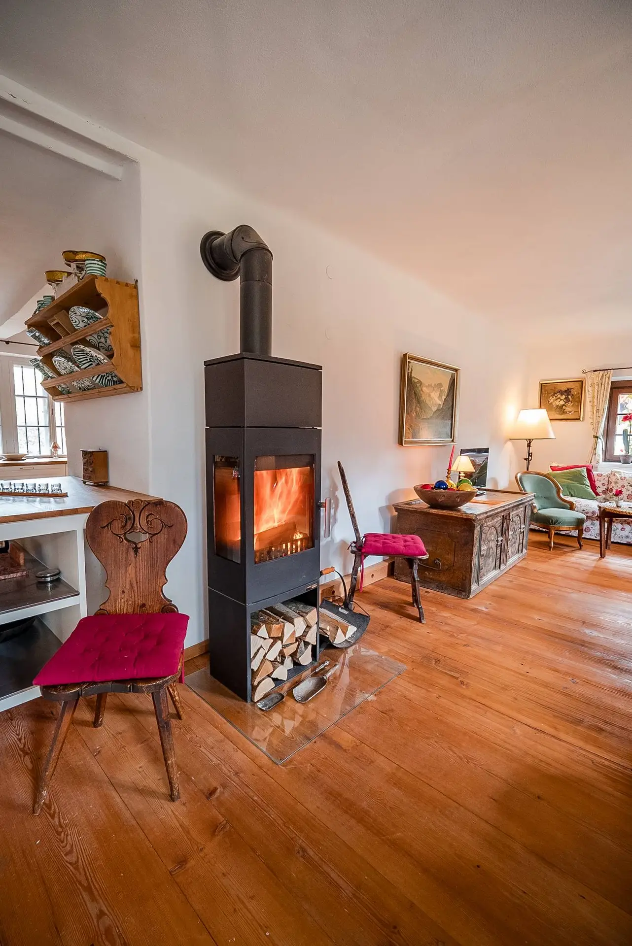 Gemütliches Wohnzimmer mit Holzofen, Brennholzlager, schwebenden Regalen, Kunstwerken und rustikalem Holzboden. Bunte Stühle sorgen für einen Farbtupfer.