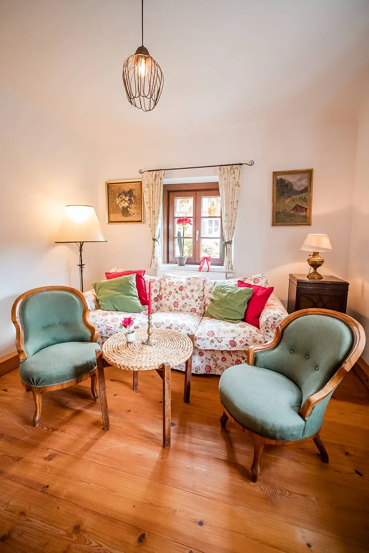Gemütliches Wohnzimmer mit zwei grünen Vintage-Sesseln, einem Sofa mit Blumenmuster und einem Holzboden. Ein Fenster im Hintergrund bietet einen Blick nach draußen. Zur Einrichtung gehören Lampen und gerahmte Gemälde von