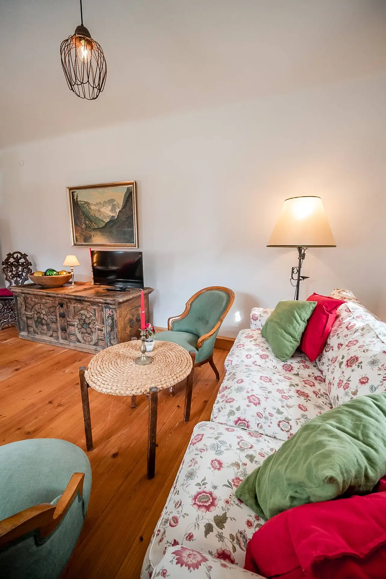 Gemütliches Wohnzimmer mit geblümtem Sofa, grünem Sessel, rundem Tisch, verzierter Holzkommode und Hängelampe.