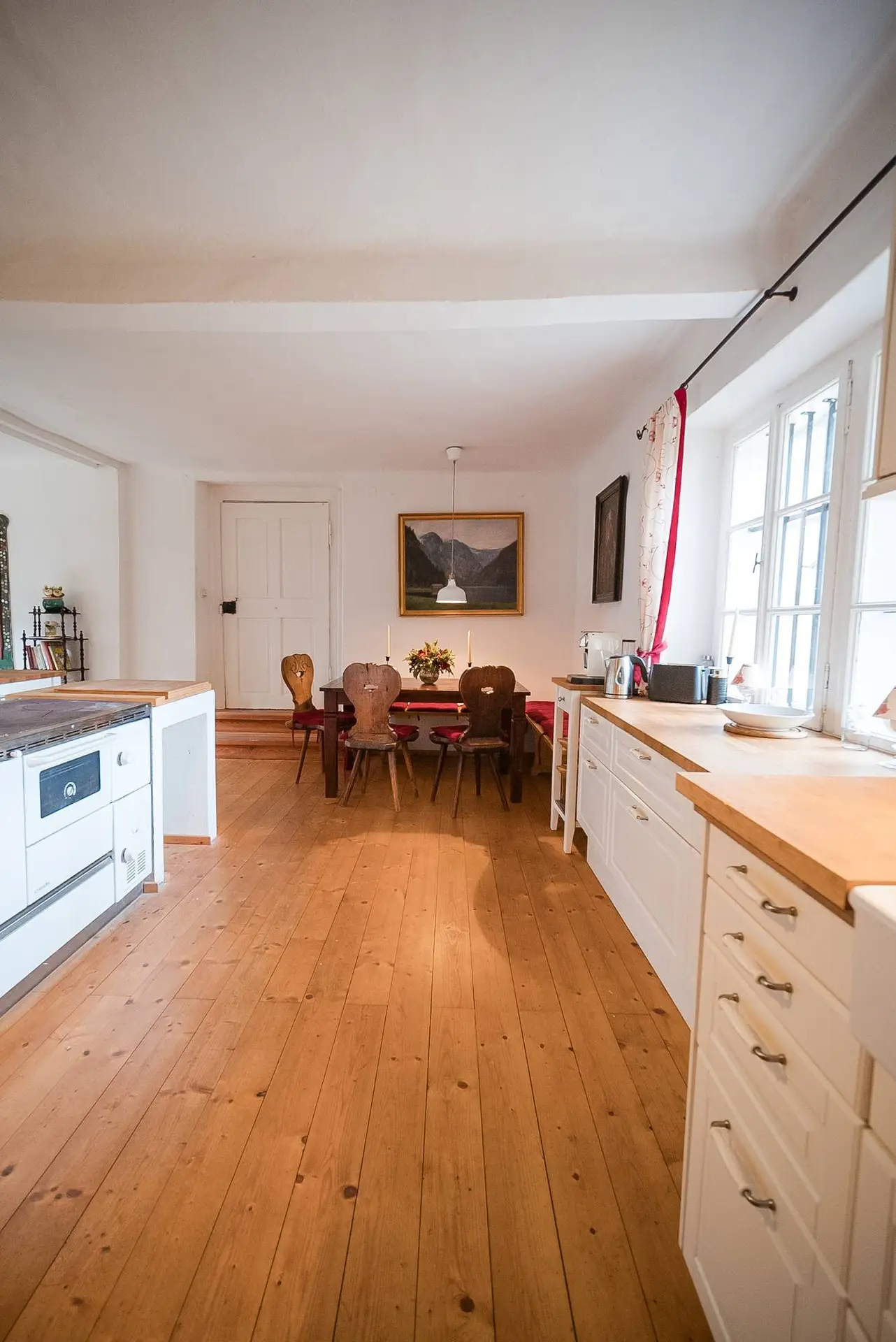 Innenraum einer gemütlichen Küche und eines Essbereichs mit Holzböden, weißen Schränken und einem Tisch für vier Personen, ergänzt durch das natürliche Licht der Fenster.