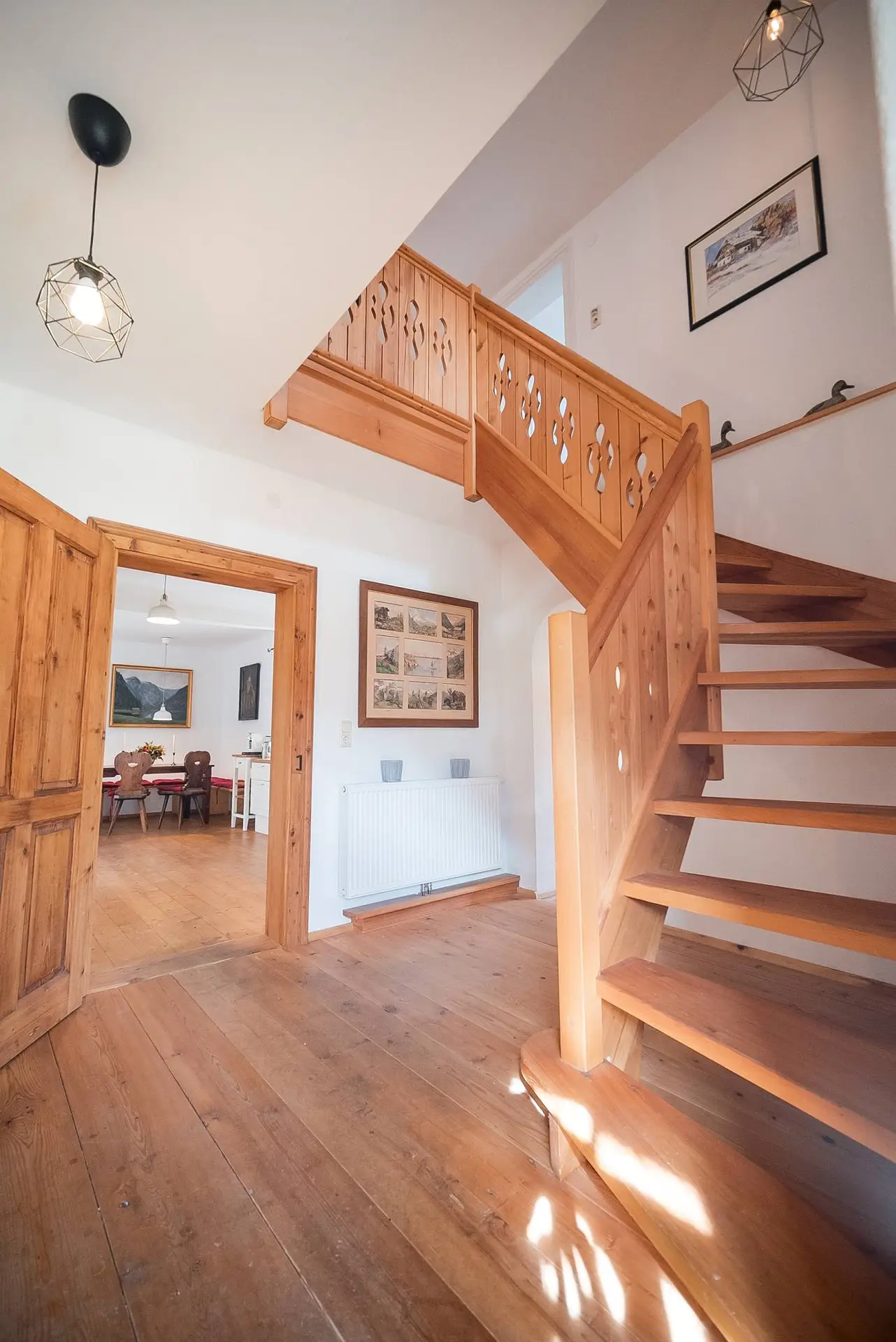 Innenansicht eines Hauses mit einer Holztreppe, die in ein oberes Stockwerk führt, und einer offenen Tür, die in ein Wohnzimmer führt.