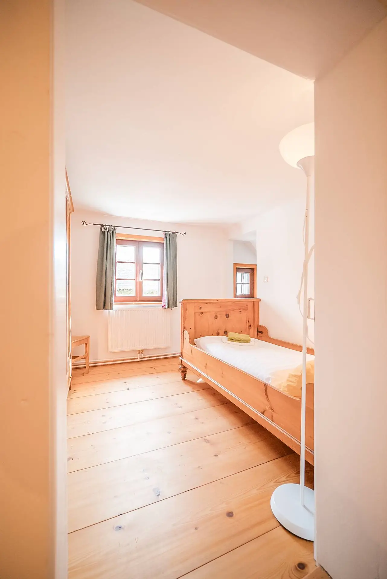 Ein helles, gemütliches Schlafzimmer mit Holzböden und -möbeln, darunter ein Bett mit weißer Bettwäsche und ein Fenster mit grauen Vorhängen.